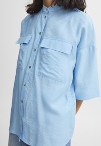 Camicia button-up azzurro chiaro con maniche corte, dotata di due taschini sul petto e colletto alla mandarino. Il materiale sembra essere strutturato e morbido.