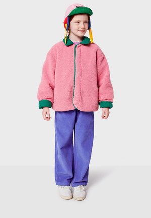 Giacca sherpa rosa con colletto verde, zip frontale e polsini a coste; abbinata a pantaloni in corduroy viola e sneakers bianche. Bambino che indossa un cappellino colorato.