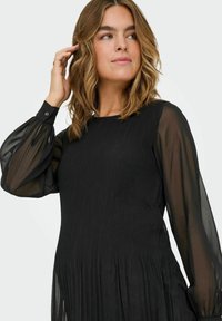 Črna obleka s rebrasto teksturo telesa in prozornimi napihnjenimi rokavi. Opremljena je z okroglim izrezom in zapenjanimi manšetami.