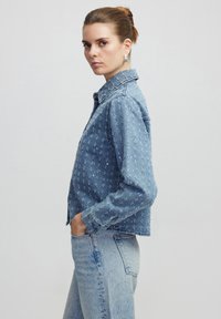 Giubbotto in denim blu chiaro con chiusura a bottoni, motivo floreale ricamato e colletto classico. Presenta una vestibilità cropped e maniche lunghe.