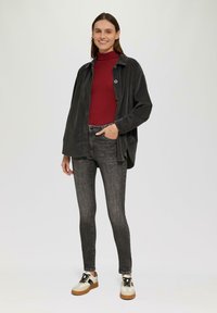Zwarte corduroy oversized jacket, rode coltrui, grijze skinny jeans en witte sneakers met zwarte accenten. Model staat tegen een neutrale achtergrond.