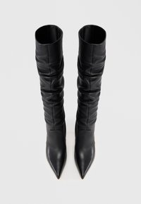 Hohe schwarze Lederstiefel mit spitzen Zehen und gerafften Details entlang des Schaftes, die auf einer weißen Oberfläche aufrecht stehen.