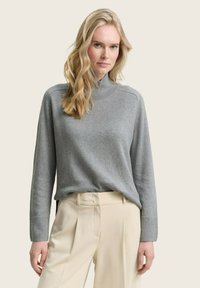 Grauer Rollkragenpullover mit langen Ärmeln und gerippten Bündchen, kombiniert mit beigefarbenen hochgeschnittenen Hosen. Glatte Textur und lockere Passform.