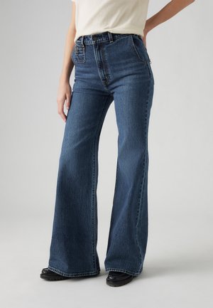 Personne portant un jean bleu taille haute à jambes larges, des chaussures noires et un haut écru rentré dans le pantalon devant un fond uni.