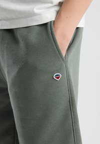 Pantaloni della tuta in cotone verde con tasche laterali, dotati di un piccolo logo ricamato a forma di triangolo in rosso, bianco e blu sulla gamba sinistra.