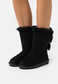 Bottines en suède noir avec un poignet en fourrure douce et deux nœuds en tissu noir à l'arrière ; dotées d'une semelle en caoutchouc texturé pour une meilleure adhérence.