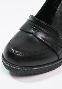 Mocassin en cuir noir avec bout arrondi, couture décorative et semelle épaisse texturée sur fond blanc.