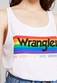 Vit linne med ett färgglatt regnbågs-mönster. Innehåller en djärv svart "Wrangler"-logotyp och horisontell text i pastellfärger nedan.