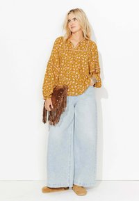 Femme blonde portant un blouse florale moutarde, un jean large clair, des chaussures slip-on beiges, tenant une pochette en franges marron contre un fond blanc.