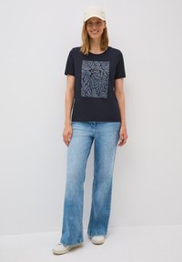 Marinblå t-shirt med ett bladmönster i grått, kombinerad med ljusblå vida jeans och vita sneakers. Stylad med en ljusfärgad keps.