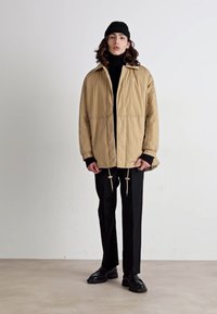 Beige gewatteerde jas met trekkoorden, gedragen over een zwart coltrui. Gestyled met zwarte broek en zwarte schoenen, tegen een neutrale achtergrond.