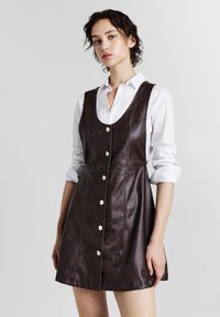 Giovane donna che indossa un vestito in pelle scura senza maniche con bottoni su una camicia bianca a maniche lunghe arrotolate.