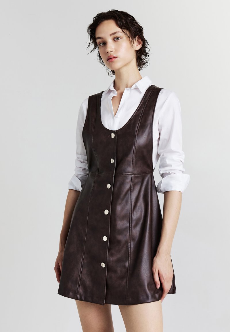 Giovane donna che indossa un vestito in pelle scura senza maniche con bottoni su una camicia bianca a maniche lunghe arrotolate.