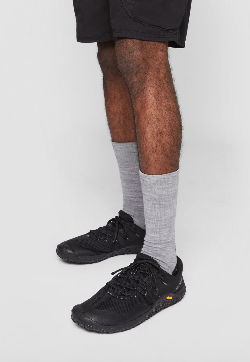 Chaussures de sport noires avec dessus en mesh texturé et fermeture à lacets. Portées avec des chaussettes grises mi-mollet. Fond blanc uni.