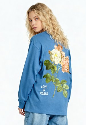 Giovane donna con lunghi capelli biondi che indossa una camicia blu con stampa floreale e la scritta "LOVE & ROSES" sulla schiena.