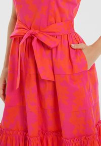 Robe rose vif avec motif floral orange, dotée d'une ceinture à la taille, d'un ourlet à volants et de poches de chaque côté. Fabriquée en tissu léger.