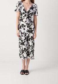 Robe florale noir et blanc avec un décolleté en V, des manches courtes bouffantes, et un lien à la taille. Tissu lisse avec une longueur midi fluide. Sandales noires à enfiler.