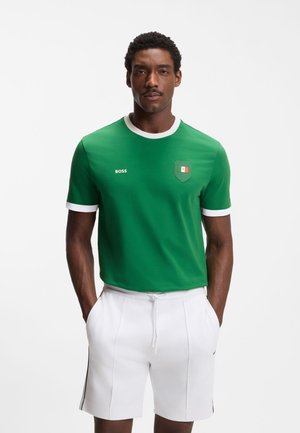 Mann trägt grünes T-Shirt mit weißem Saum und Mexiko-Flaggen-Aufnäher, kombiniert mit weißen Shorts mit Kordelzug, steht mit den Händen in den Taschen.