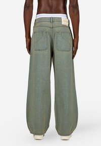 Plati kāju džinsi maigas zaļas krāsas, ar teksturētu virsmu, divām aizmugurējām kabatām un baltu jostas vietu.