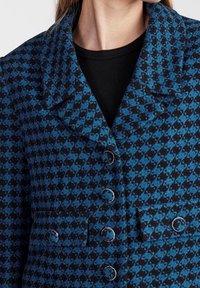 Modrý a černý kostkovaný blejzr se strukturovaným límcem, šesti kulatými knoflíky a boční kapsou, který předvádí texturovaný vzor látky.