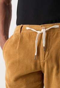 Bruine corduroy broek met een textuur op het oppervlak, voorzien van een knoopsluiting en een witte trekkoorden in de taille. Zijzak zichtbaar.