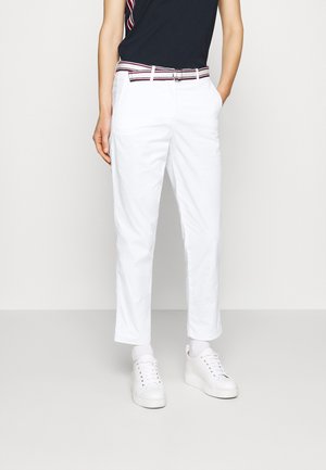 Person iført hvide cropped bukser, hvide sneakers, en sort top og et stribet bælte med hænderne i lommerne mod en ensfarvet baggrund.