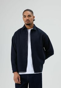 Donkerblauw overhemd met knopen, gemaakt van zachte stof, met een kraag en lange mouwen, gedragen over een wit T-shirt met ronde hals. Donkere spijkerbroek.