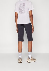 Grå shorts, knälånga, svart t-shirt med berg- och ormbunksgrafik på ryggen, vita strumpor med grå mönster, beige sneakers.