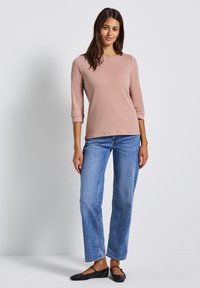 Frau trägt ein hellrosa Langarm-Oberteil, blaue gerade Jeans und schwarze Mary Jane Ballerinas und steht vor einem weißen Hintergrund.