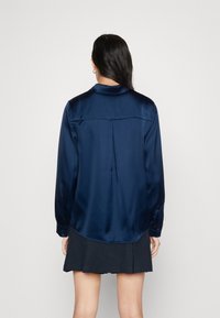 Kvinde med langt mørkt hår iført en navy blå satin-langærmet skjorte og en sort nederdel, stående med ansigtet mod en hvid baggrund.