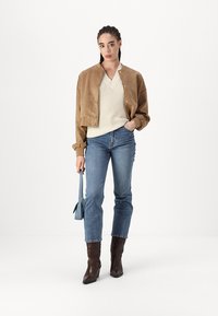 Une veste bomber en daim brun portée sur un pull en tricot crème, associée à un jean droit bleu et à des bottes à talons marron. Tient un sac bleu clair.