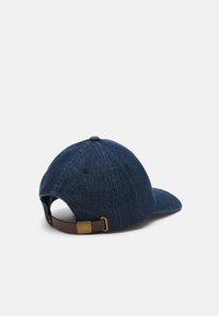 Gorra de béisbol de mezclilla en azul oscuro, con visera curvada, tejido texturizado y una correa marrón con una hebilla de metal dorado en la parte trasera.
