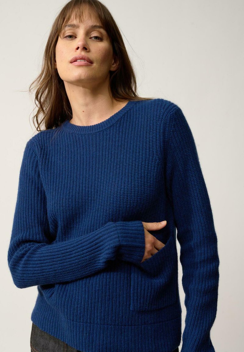 Pull en tricot bleu avec texture côtelée, manches longues, col rond et une poche à l'avant. Le tissu semble chaud et décontracté.