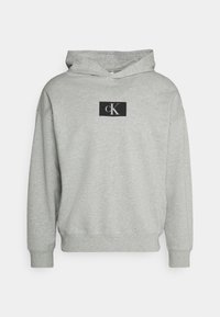Felpa grigia in cotone con una toppa nera frontale che presenta un logo "cK" bianco, maniche lunghe e cappuccio con coulisse. Design morbido e casual.