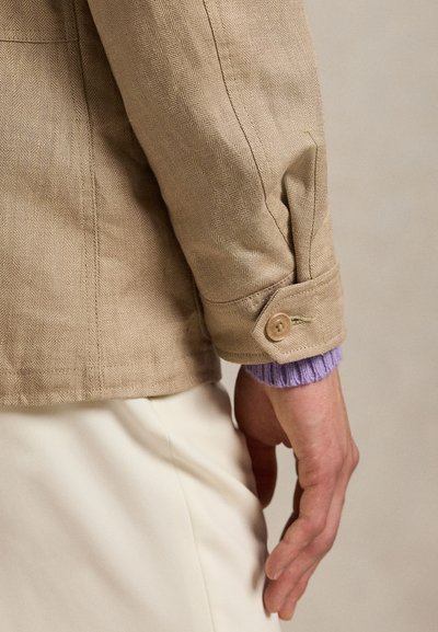 Person's hand resting by side wearing beige textured jacket with buttoned cuff over lavender sweater and white pants.