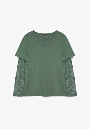 Grünes Langarmoberteil mit einfarbigen kurzen Ärmeln, das über transparenten floralen Spitzenärmeln und passendem Spitzen-V-Ausschnitt eingesetztem Stoff getragen wird.