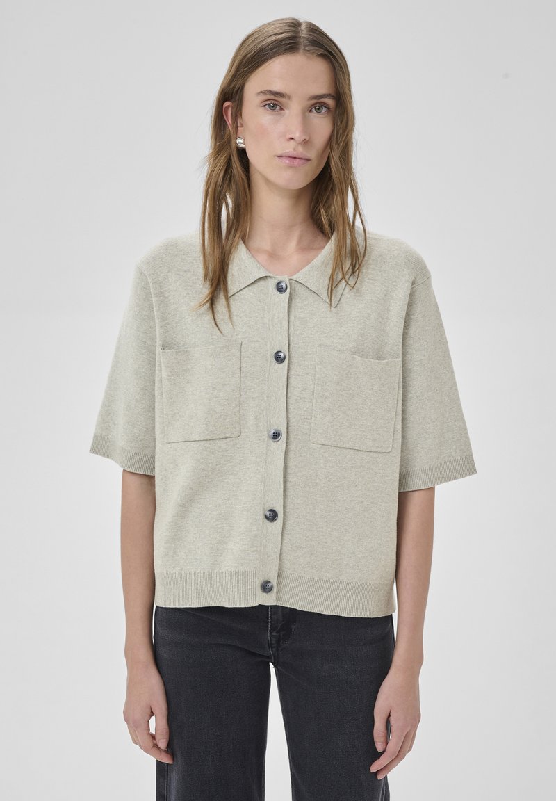Femme portant un cardigan beige à manches courtes boutonné avec deux poches avant et un pantalon noir, debout devant un fond uni.