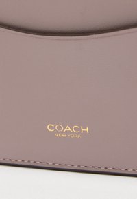 Tuva ādas tuvinājums violeti rozā krāsā ar zelta "Coach New York" logotipu un detalizētu šuvumu apakšējā malā.