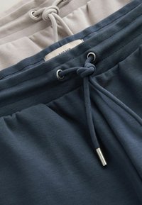 Gros plan sur des ceintures de pantalons de survêtement bleu marine et beige, avec des cordons de serrage noués en nœuds simples et des embouts métalliques.