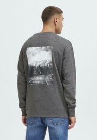 Mann som vender bort med en grå hettegenser med et svart-hvitt trykk av fjell- og skogsvei på baksiden, kombinert med blå jeans.