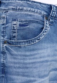 Vaqueros de mezclilla azul con un diseño clásico, que cuentan con un bolsillo frontal con costuras en contraste y detalles de remaches metálicos, textura suave y lavado desgastado.