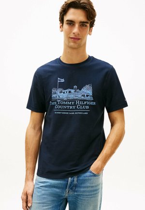 Joven con una camiseta azul marino con un dibujo lineal de un club de campo y el texto "The Tommy Hilfiger Country Club" y vaqueros azul claro.