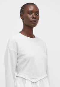 Sudadera gris clara con mangas largas y un escote redondo, que presenta un dobladillo inferior blanco fruncido y una textura suave.