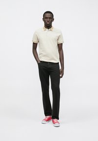 Beige polo skjorte med gul krage, sammen med svarte jeans og røde sneakers. Modellen står rett opp mot en enkel bakgrunn.