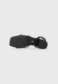 Sandale à talons hauts noirs avec un orteil rectangulaire lisse et une semelle texturée. Elle possède des straps entrecroisés et une fine lanière à la cheville pour le soutien.