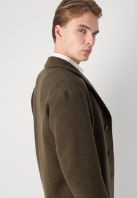 Brun ullblandad kappa med kiltkrage, dubbelknäppta knappar, sidofickor och en slät textur. Visad på en modell från sidan.