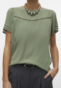 Grøn bluse med korte ærmer, detaljer med blondekant og en glat overflade, parret med mørke bukser og en stor metallic perlekæde.