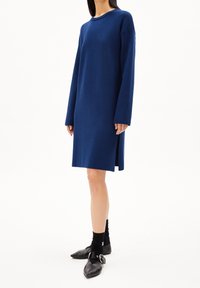 Robe bleu marine à manches longues en tissu texturé, avec un col rond et des fentes latérales, associée à des chaussures pointues noires et des chaussettes.