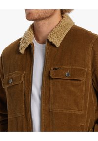 Ruskea corduroy-takki, jossa on vaaleanruskea fleece-kaulus. Takissa on kaksi etutaskua ja vetoketjusuljenta, ja sen kangas on teksturoitu pystysuorilla urilla.