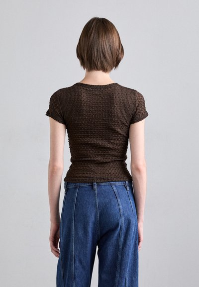 Hnědý textured top s krátkým rukávem a přiléhavým střihem, v kombinaci s modrými denimovými kalhotami s vysokým pasem, které mají výrazné švy a volný siluetu.